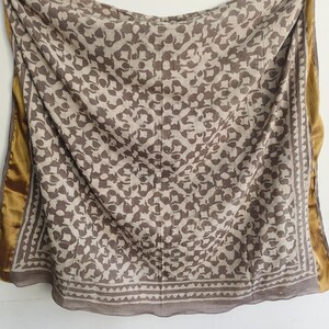 Puede incluir: Bufanda beige y marr&oacute;n estampada con un dise&ntilde;o floral repetido. La bufanda tiene un borde dorado y un borde geom&eacute;trico a lo largo de los bordes. La tela est&aacute; drapeada, mostrando la textura y el patr&oacute;n de la bufanda.