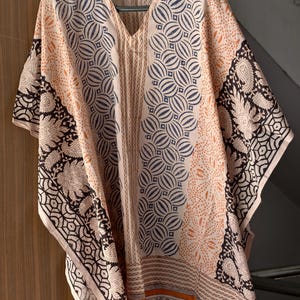 May include: A beige, V-neck kaftan with a geometric and paisley print. The kaftan features a central panel with blue and white circular patterns, bordered by panels with black and white paisley and geometric designs. The sleeves are short and wide.