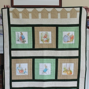 May include: A beige and green patchwork quilt with a Peter Rabbit theme. The quilt features nine squares with different Peter Rabbit illustrations. The quilt is edged with a brown and beige trim.