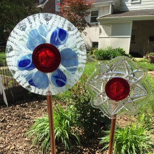 May include: Two glass flower decorations with red centers and copper stems. The flowers are made from vintage glass plates with a clear, textured design. The flowers are displayed in a garden setting.