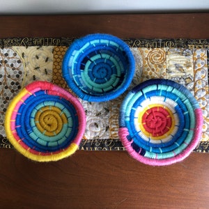 以下が含まれることがあります： 円形のデザインが施された、カラフルな織りバスケット3つ。バスケットは、青、黄、ピンク、白の糸でできています。バスケットは、模様のある表面に一列に並べてあります。
