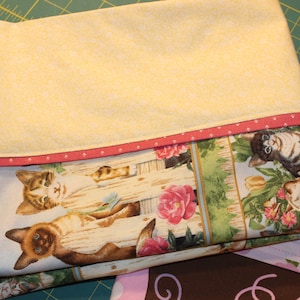 May include: A yellow fabric with a pink and white polka dot trim. The fabric is folded over a blue fabric with a print of cats sitting on a fence with flowers.