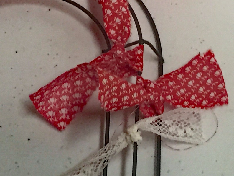 May include: Red and white patterned fabric ribbon tied around a metal wreath form. The ribbon is tied in a bow and the ends are frayed.