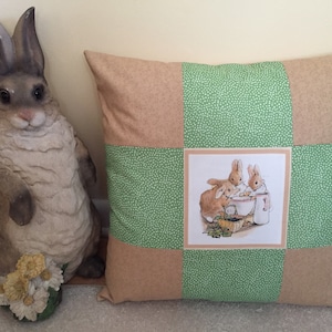 May include: A green and brown patchwork pillow with a square image of three bunnies in the center. The bunnies are brown and white and are eating from a bowl. The pillow is decorated with white polka dots.