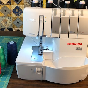 May include: A white Bernina L 450 overlock sewing machine with four spools of thread in various colors. The machine has a clear plastic cover over the needle area and a white plastic tray on the right side.