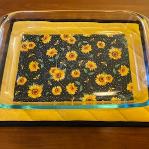 Puede incluir: Una fuente de horno de vidrio transparente con asas rojas descansa sobre un salvamanteles acolchado amarillo con borde negro. Dentro de la fuente hay una tela negra con un estampado de girasoles.