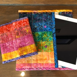 Op de afbeelding: Een regenboogkleurige, gewatteerde tablethoes met een kleinere bijpassende hoes. De grotere hoes heeft een tablet erin. De hoes is gemaakt van stof met een bloemenpatroon.