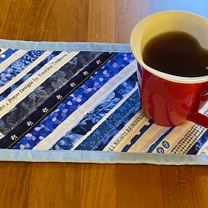 May include: A blue and white quilted coaster with the text "Joy Wing and a Prayer Designs for Timeless Treasures P" and "ALL RIGHTS RESERVED" on it. A red mug with a beige rim and brown liquid inside is sitting on the coaster.