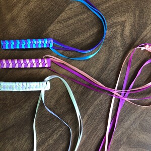 May include: Three braided ribbon hair ties with long tails. The hair ties are in shades of purple, pink, and white. The tails are in matching colors.