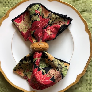 May include: A black and red floral napkin with a gold and brown wooden napkin ring. The napkin is folded and placed on a white plate with a gold rim.