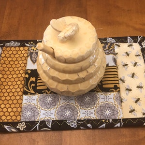 May include: A ceramic honey pot with a bee-shaped lid sits on a rectangular quilted mat. The mat features a patchwork design with honeycombs, floral patterns, and bee motifs in shades of gold, black, and cream. The edges are trimmed with a decorative stitch.
