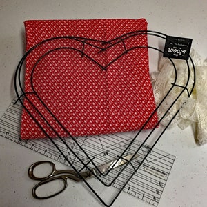 May include: A red fabric with white polka dots, a pair of scissors, a ruler, a white lace ribbon, and two black metal heart-shaped hoops. The hoops are stacked on top of each other. The fabric is laying on top of the ruler. The ribbon is laying on top of the hoops. The text on the tag attached to the hoops reads "The Bloom Room".