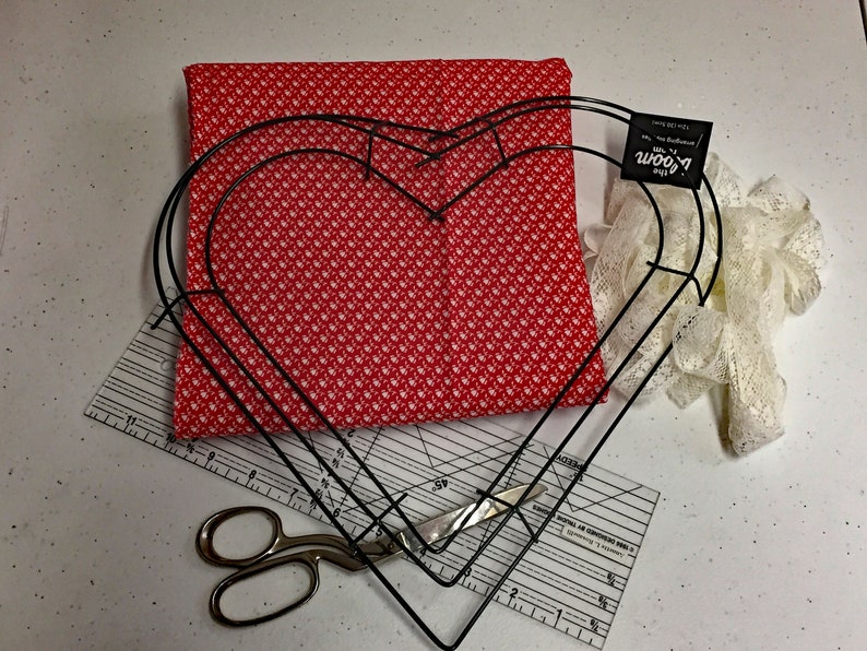 May include: A red fabric with white polka dots, a pair of scissors, a ruler, a white lace ribbon, and two black metal heart-shaped hoops. The hoops are stacked on top of each other. The fabric is laying on top of the ruler. The ribbon is laying on top of the hoops. The text on the tag attached to the hoops reads "The Bloom Room".