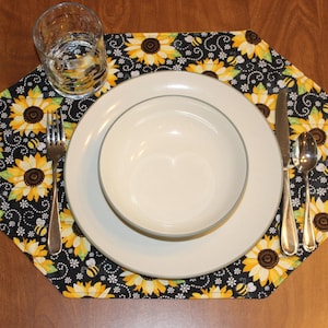 May include: A black and yellow sunflower placemat with a white plate, bowl, fork, knife, and spoon on top.