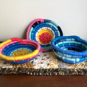 May include: Three small, round, coiled rope bowls. One bowl features a vibrant yellow center with pink, blue, and yellow outer rings. Another bowl displays a bright yellow center with pink and blue rings. The third bowl is shades of blue.
