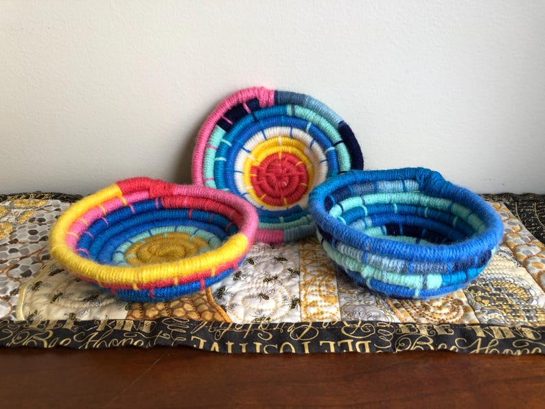 May include: Three small, round, coiled rope bowls. One bowl features a vibrant yellow center with pink, blue, and yellow outer rings. Another bowl displays a bright yellow center with pink and blue rings. The third bowl is shades of blue.