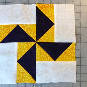 May include: A quilt block with a pinwheel design. The block is made of four triangles of purple fabric and four triangles of yellow fabric with a white floral pattern. The triangles are arranged in a pinwheel pattern, with the purple triangles pointing towards the center of the block.