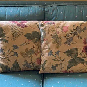 May include: Two square throw pillows with a floral pattern. The pillows feature a beige background with a repeating design of pink, purple, and blue flowers, along with green leaves and stems. The pillows are placed on a teal couch.