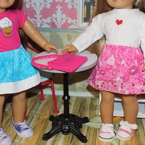 Puede incluir: Dos muñecas con vestidos coloridos y zapatillas de deporte están cerca de una mesa pequeña con un bolso rosa. La muñeca de la izquierda lleva una camisa rosa con un diseño de cupcake y una falda azul y blanca. La muñeca de la derecha lleva una camisa blanca con un corazón rojo y una falda floral rosa.