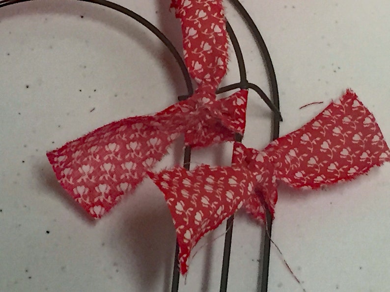 May include: Red and white fabric ribbon tied around a metal wreath form. The ribbon has a small white floral pattern.