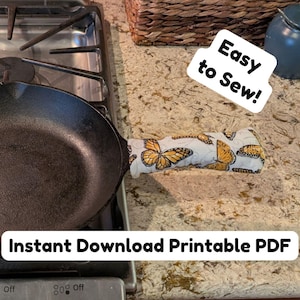 May include: A black cast iron skillet with a fabric handle cover featuring a yellow and black butterfly pattern. The image includes the text Easy to Sew! and Instant Download Printable PDF. The skillet is positioned on a stovetop surface.