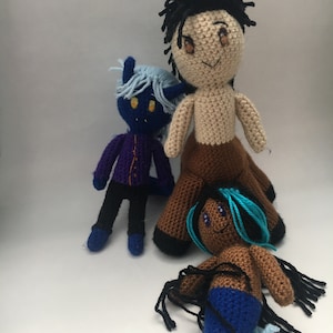 May include: Three crocheted plush toys: a blue and purple demon, a brown-skinned mermaid with turquoise hair, and a brown-skinned person with black hair and a brown skirt.