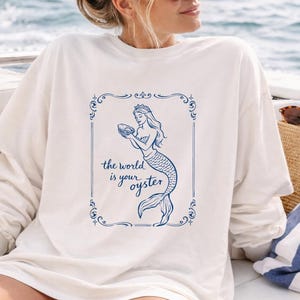 Peut inclure: Sweat-shirt crème avec une illustration bleue d'une sirène tenant une huître, encadrée par une bordure décorative. Le texte "the world is your oyster" est en dessous. Le sweat-shirt a des manches longues.