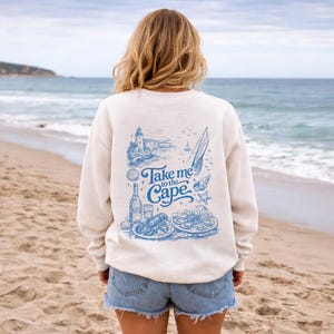 May include: A cream-coloured sweatshirt featuring a blue graphic with the text "Take me to the Cape." The design includes a lighthouse, sailboat, and food illustrations. The person is wearing denim shorts on a sandy beach.