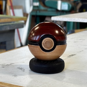 May include: A wooden Poké Ball replica, featuring a two-tone design with a dark brown top and a light brown base. The ball has a black band and a circular wooden button. It sits on a small, black, circular base, showcasing detailed craftsmanship.