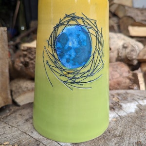 May include: A yellow and green ceramic mug with a blue circle inside a black wire design.