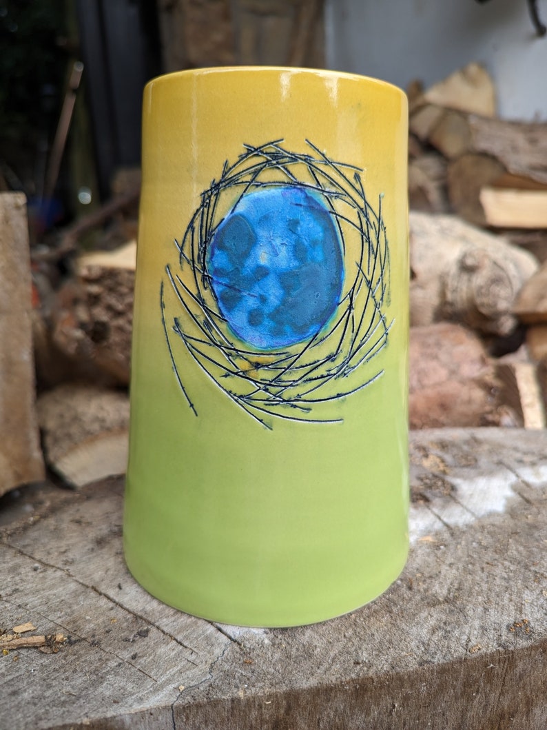 May include: A yellow and green ceramic mug with a blue circle inside a black wire design.
