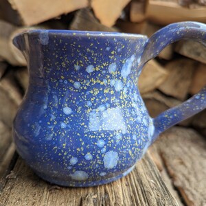 May include: A blue ceramic jug with a speckled glaze in shades of blue, white, and gold. The jug has a rounded body and a handle.