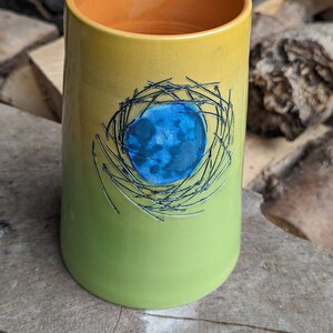 May include: A green and yellow ceramic mug with a blue and black design of a bird's nest.