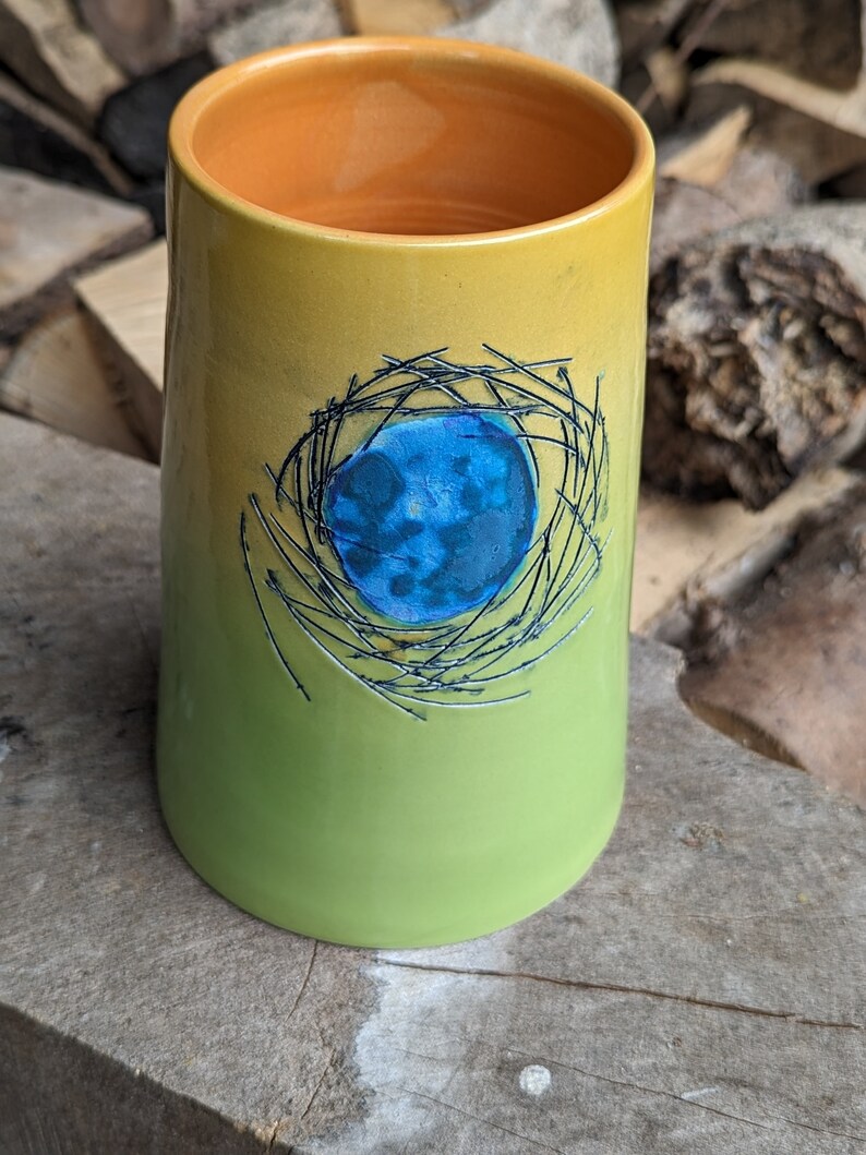 May include: A green and yellow ceramic mug with a blue and black design of a bird's nest.