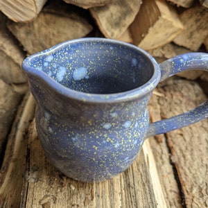 May include: A small blue ceramic creamer with a speckled glaze. The creamer has a rounded body and a handle.
