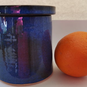 May include: A blue ceramic jar with a lid, next to an orange. The jar has a glossy finish and a slight iridescent sheen.