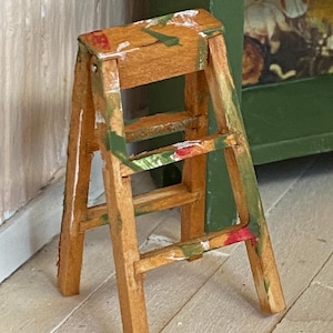 May include: A small, wooden step stool with a natural wood finish, accented with green, red, and white paint. The stool has two steps and a folding design. The legs are angled for stability. The overall style is rustic and handmade.