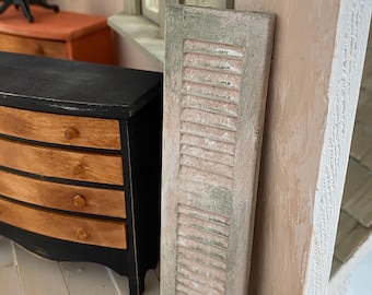 May include: A miniature, weathered, rectangular shutter with horizontal slats. The shutter is painted in a distressed, muted green and cream color scheme, suggesting a vintage aesthetic. It is leaning against a wall, next to a miniature chest of drawers.