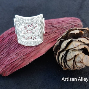 Peut inclure: Une bague en filigrane argentée avec un motif carré. La bague est posée sur une surface texturée rouge et brune. Artisan Alley est écrit en blanc en bas de l'image.