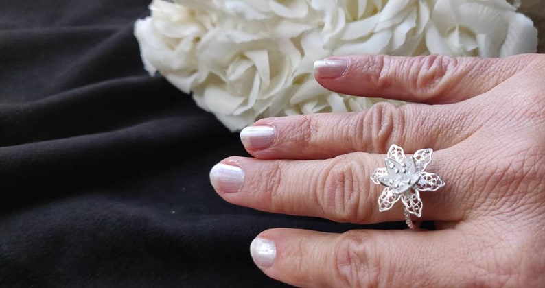 Pode incluir: Um anel de filigrana de prata com um design de flor. O anel est&aacute; em um dedo com esmalte de unha branco.