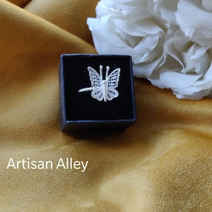 May include: A silver butterfly ring in a black ring box. The ring is delicate and intricate, with a filigree design. The box is on a yellow fabric surface.
