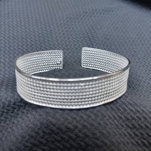 Op de afbeelding: Een zilverkleurige manchetarmband met een geometrisch openwerkontwerp. De armband heeft een rechthoekige vorm met een reeks kleine, gelijkmatig verdeelde gaten. Het open ontwerp laat licht door, waardoor een delicate uitstraling ontstaat. De armband is waarschijnlijk van metaal.