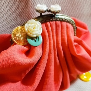 May include: A coral colored fabric coin purse with a metal clasp and decorative buttons. The buttons are white, gold, and green.