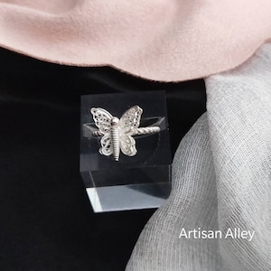 May include: A silver butterfly ring with intricate details. The ring is displayed on a clear acrylic stand.