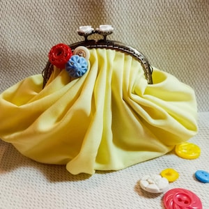 Może przedstawiać: Żółta skórzana torebka na monety z metalową ramką i kolorowymi guzikami. Torebka jest otwarta i leży na fakturowanej powierzchni z rozrzuconymi wokół niej guzikami.