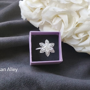 May include: A silver filigree flower ring in a purple box. The ring is delicate and intricate, with a floral design. The box is small and square, with a purple velvet lining.