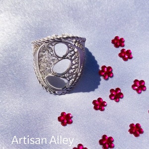 May include: A silver filigree ring with a large oval cutout in the center. The ring is on a white background with pink flower-shaped embellishments.