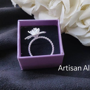Pode incluir: Um anel de prata com um design de flor em uma caixa roxa. A caixa est&aacute; sobre uma superf&iacute;cie preta. Artisan Alley.
