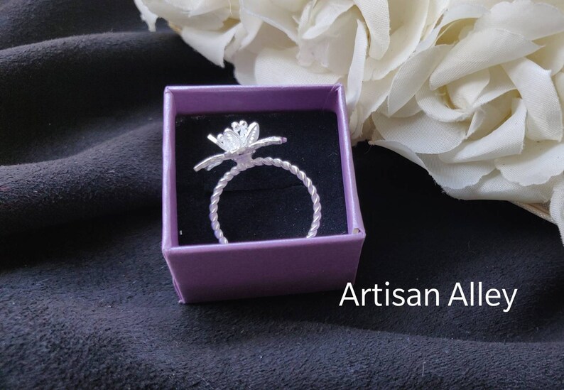 Pode incluir: Um anel de prata com um design de flor em uma caixa roxa. A caixa est&aacute; sobre uma superf&iacute;cie preta. Artisan Alley.
