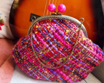 May include: A vibrant, textured clutch bag with a kiss-lock closure and a chain strap. The bag is crafted from woven fabric in shades of pink, red, purple, blue, and yellow. The clasp has two pink, spherical embellishments.